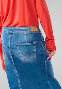 Jupe en denim bleu clair à taille haute, avec une poche arrière dotée de coutures arrondies et une étiquette de marque beige. Associée à un haut à manches longues orange.