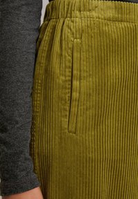 Olijfgroene corduroy broek met verticale ribbels, elastische tailleband en zijzakken. Ernaast is een grijze lange mouw top zichtbaar.