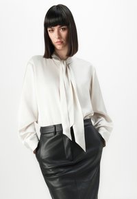 Femme avec des cheveux noirs lisses portant une blouse blanche avec un nœud au cou et une jupe en cuir noir taille haute, les mains dans les poches.