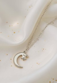 Srebrny naszyjnik z wisiorkiem w kształcie półksiężyca i gwiazdy, na delikatnym łańcuszku, wyeksponowany na miękkiej białej tkaninie z złotymi akcentami gwiazd.