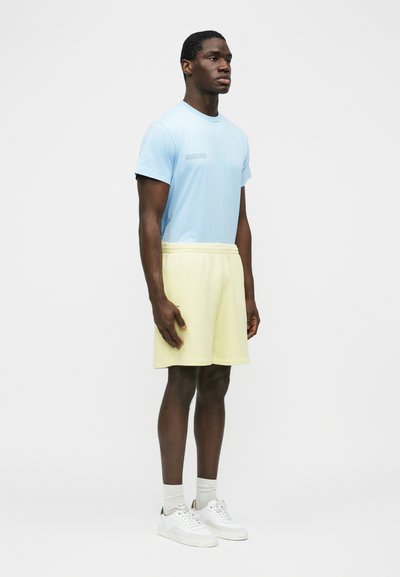 Young man standing in light blue T-shirt, pale yellow shorts, white socks, and white sneakers against a plain white background.