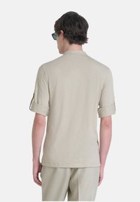Camicia beige a maniche corte con vestibilità rilassata, caratterizzata da maniche arrotolate e una texture liscia, vista da dietro su uno sfondo semplice.