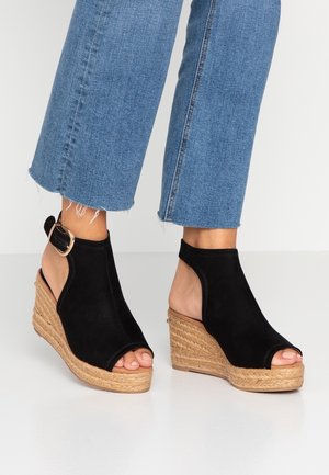 Sorte ruskinds wedgesandaler med åben tå og slingback-design. Juteindhyllet kilehæl for tekstur og kontrast. Denim cropped bukser synlige.
