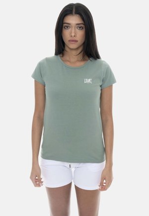 Femme portant un T-shirt vert sauge à manches courtes avec le logo "LEONE" et un short blanc, debout face à un fond uni.