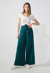 Pantalon large teal avec une taille nouée et un tissu texturé, associé à un haut blanc ajusté et des baskets blanches. Le modèle pose avec une main sur la hanche.