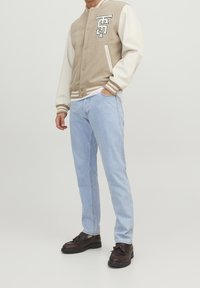 Beige varsityjacka med vita ärmar, randig trim och en patch. Ljusblå jeans och mörka skor med fransdetalj kompletterar outfiten.