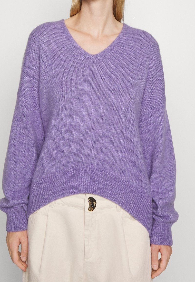 Maglione a V in maglia viola con maniche oversize e orlo a coste, abbinato a pantaloni chiari con bottone decorativo.