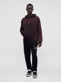 Homme debout portant un sweat à capuche bordeaux foncé avec poche avant et un pantalon de jogging noir avec un détail graphique ainsi que des baskets noires et blanches.