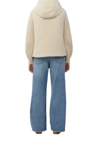 Person in beigem Kapuzen-Sherpa-Weste über einem gestrickten Pullover und hellblauen weit geschnittenen Jeans, steht mit dem Rücken zur Kamera vor einem weißen Hintergrund.