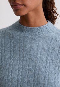 Helles blaues gestricktes Sweater mit hohem Kragen, der ein strukturiertes Zopfmuster und ein weiches, flauschiges Material aufweist. Nahaufnahme des Ausschnitts.