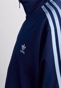 Marinblå Adidas träningsjacka med ljusblå ränder, hög krage och subtil branding på bröstet. Slät tygtextur. Dragkedjestängning.