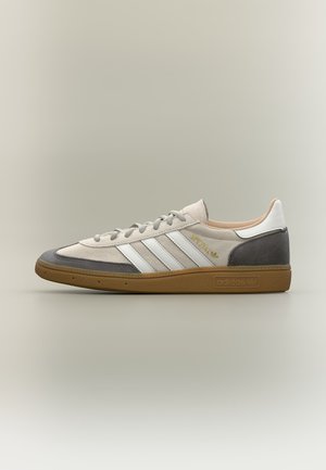 Niski profil tenisówki Adidas z białymi paskami, beżową cholewką, szarymi akcentami na palcach i pięcie oraz gumową podeszwą, widziane z boku.