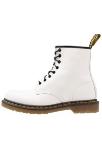 Stivaletto bianco in pelle con lacci neri, cuciture gialle e un cinturino per il tallone. Texture liscia con una forma della punta arrotondata.