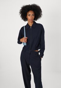 Giacca cropped a righe navy con zip frontale e colletto, abbinata a pantaloni a vita alta coordinati. Accentuata da una tracolla blu chiaro.