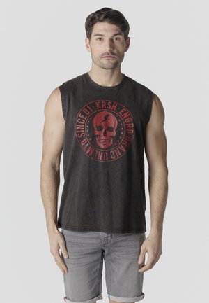 Hombre con camiseta sin mangas negra con gráfico de calavera roja y pantalones cortos grises remangados, frente a un fondo claro y liso.