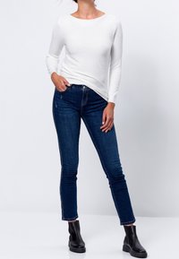 Top lungo a maniche lunghe bianco a coste abbinato a jeans skinny blu scuro e stivaletti neri. L'outfit enfatizza una silhouette aderente.