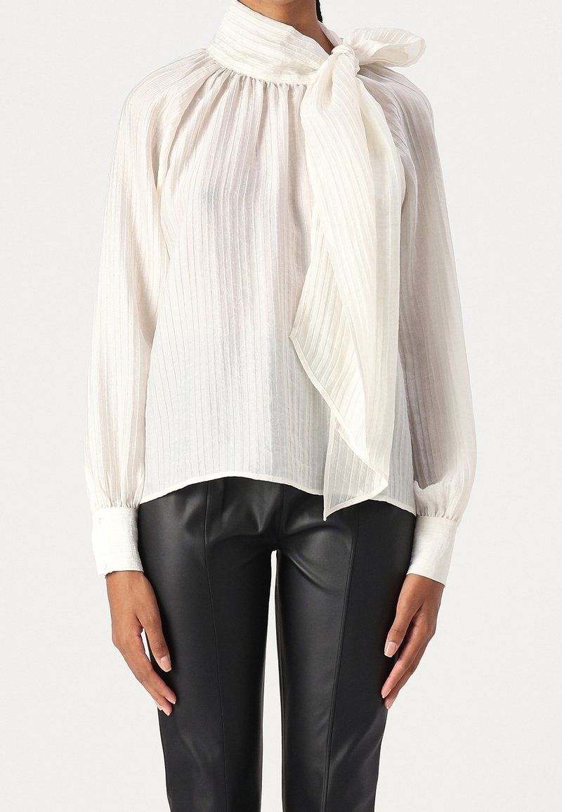 Personne portant une blouse blanche transparente à manches longues avec des rayures verticales et un grand nœud au cou, associée à un pantalon en cuir noir.