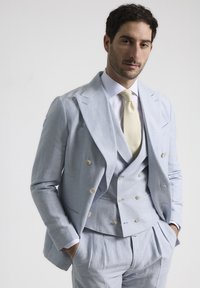 Traje de lino azul claro de doble botonadura con chaleco, con solapas de muesca y botones blancos; combinado con una corbata crema sobre una camisa blanca.
