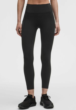 Frau trägt schwarze Leggings mit hohem Bund und Seitentaschen sowie schwarze Sportschuhe, steht vor einem einfarbigen hellen Hintergrund.