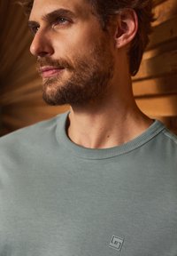 Harmaa lyhythihainen collegepaita, jossa on pyöreä pääntie ja hienovarainen logo. Pehmeä materiaali ja rento muotoilu lämpimän sävyisessä taustassa.