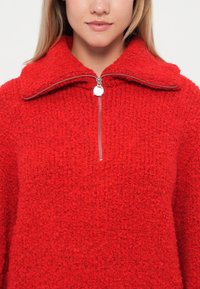 Rode, gestructureerde pullover met een hoge kraag en zilveren rits. De stof lijkt zacht en pluizig.