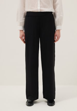 Personne portant un pantalon noir ample, des chaussures noires et un chemisier blanc transparent à manches longues, debout devant un fond uni.