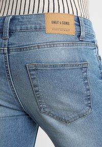 Jeans de mezclilla azul con un acabado desgastado, que presentan una etiqueta de cuero marrón claro con la inscripción "ONLY & SONS" y un diseño de un solo bolsillo trasero.