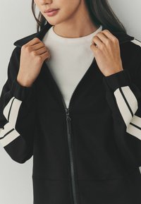 Chaqueta negra con cremallera y rayas blancas en las mangas, cuello de canalé y tejido texturizado. El modelo sostiene la cremallera con ambas manos.