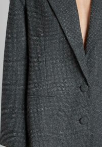 Grijze blazer met notch lapel, dubbele knopen en een visgraatstructuur met subtiele glans. Inclusief een zijzak.