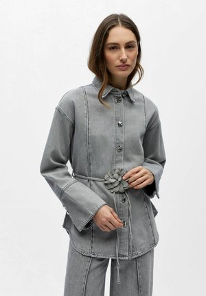 Femme portant une veste en jean grise à manches larges et une ceinture florale, associée à un pantalon en jean gris assorti, debout devant un fond blanc.