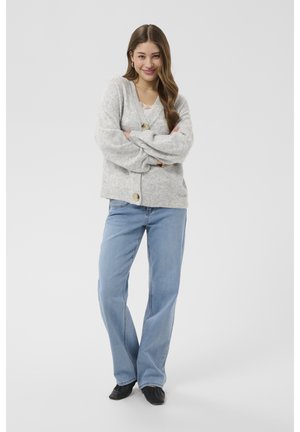 Mlada ženska stoji z rokami sklenjenimi, oblečena v svetlo sivi kardigan, belo majico, svetlo modre kavbojke in črne čevlje, na enobarvnem ozadju.