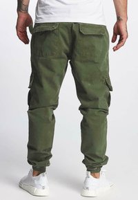 Zielone spodnie cargo z elastycznymi mankietami, tylnymi kieszeniami i bocznymi kieszeniami cargo, w połączeniu z białymi sneakersami na jednolitym tle.