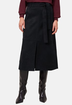 Femme portant une jupe midi en suede noire avec une fente à l'avant et un lien à la taille, assortie à des bottes noires à bout pointu arrivant jusqu'aux genoux et un pull bordeaux.