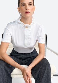 Top bianco a maniche corte con colletto arricciato e dettaglio centrale con bottone, abbinato a pantaloni scuri con texture. Tessuto liscio e design strutturato.