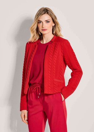 Vrouw met blond haar die een rode textuur jas, een rode top en rode broek draagt, staand met één hand in de zak tegen een neutrale achtergrond.