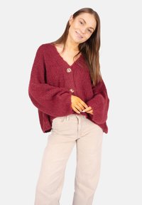 Cardigan en tricot bordeaux avec des manches surdimensionnées, un col en V et trois boutons décoratifs. Associé à un pantalon large beige clair.