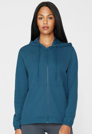 Frau mit langen dunklen Haaren, die einen türkisfarbenen Hoodie mit Reißverschluss, Fronttaschen und Kordeln trägt, steht vor einem schlichten weißen Hintergrund.