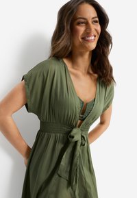 Φόρεμα σε ελαιί πράσινο με κοντά μανίκια, ελαστική μέση με εφέ σφηκοφωλιάς και λεπτομέρεια ζώνης, με βαθύ V-λαιμόκοψη και αέρινη  υφή.