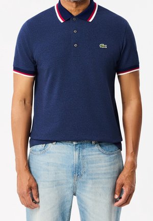 Mann trägt ein marineblaues Poloshirt mit rot-weiß-blau gestreiftem Kragen und Ärmeln, kombiniert mit hellblauen Jeans.