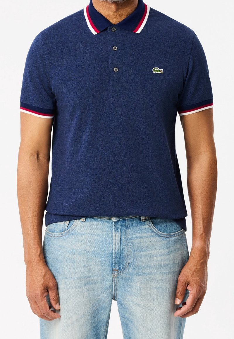Homme portant un polo bleu marine avec un col et des manches rayés rouge, blanc et bleu, associé à un jean en denim bleu clair.