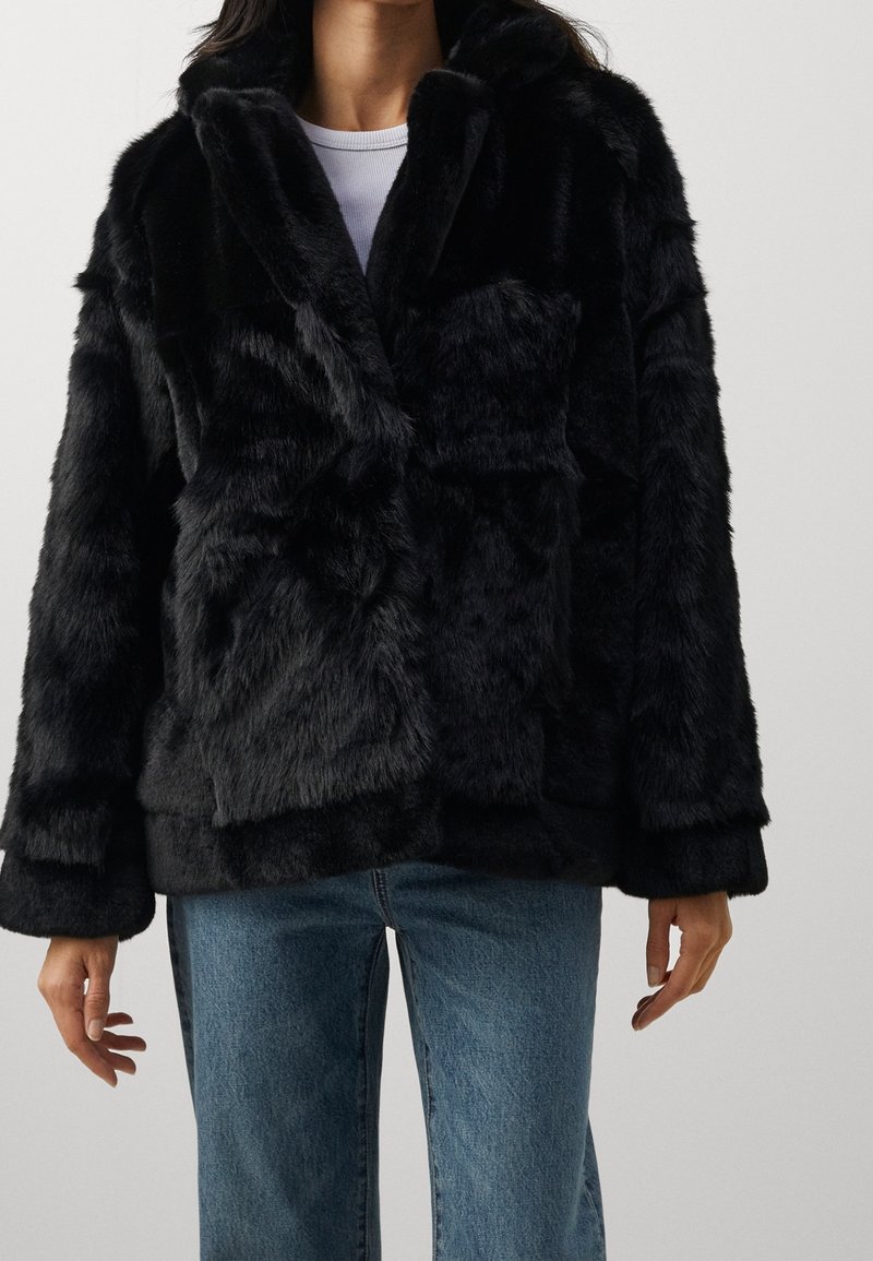 Personne portant une veste en fausse fourrure noire par-dessus une chemise blanche et un jean en denim bleu, debout les bras détendus le long du corps.