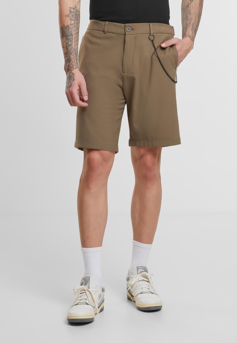 Shorts marron à coupe décontractée, fermeture à bouton unique, deux poches avant, et un détail en chaîne sur le côté droit. Portés avec des chaussettes blanches et des baskets.