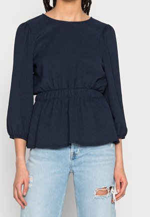 Top peplum blu navy con maniche puff a tre quarti, vita arricciata e tessuto testurizzato, abbinato a jeans strappati blu chiaro.