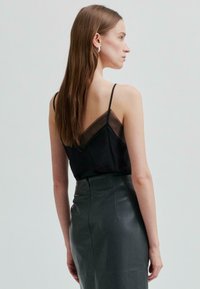 Nainen, jolla on pitkät ruskeat hiukset, käyttää hopeisia renkaita, mustaa läpikuultavaa toppia, jossa on mustat pitsireunat, sekä korkeavyötäröisiä tummasta nahasta tehtyjä hameita, käännettynä pois päin.