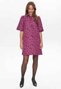 Kvinna som ler, iklädd en kortärmad, kragklädd, lila blommig texturdress, svarta genomskinliga strumpbyxor och svarta spetsiga klackar, stående framför vit bakgrund.