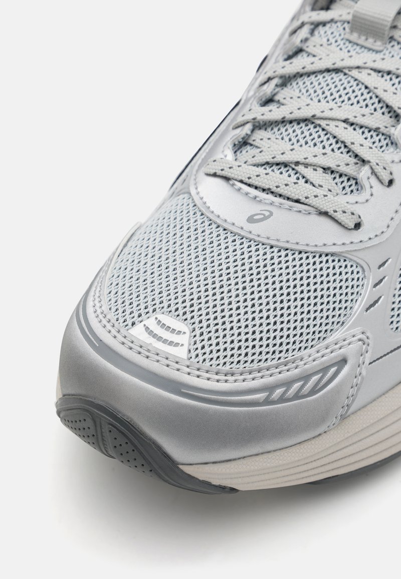 Chaussure de course grise avec une tige en mesh, des lacets plats et une semelle texturée. Les détails notables incluent une finition brillante et un logo discret.