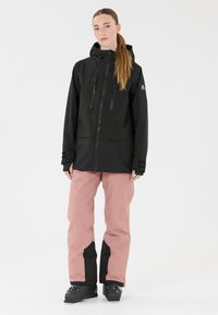 Schwarze wasserdichte Jacke mit Kapuze, Frontreißverschlüssen und zwei Brusttaschen. Kombiniert mit rosa Hosen mit schwarzen Akzenten und schwarzen Stiefeln.