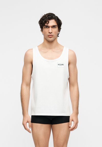 Νέος άνδρας με σκούρα σγουρά μαλλιά φοράει λευκό αμάνικο Moschino Active μπλουζάκι και μαύρα εφαρμοστά σορτς, στέκεται μπροστά σε απλό φόντο.