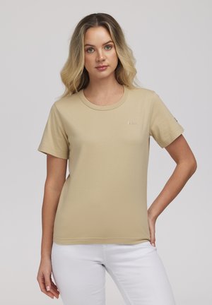 Kobieta o blond włosach, ubrana w beżowy t-shirt z krótkim rękawem i białe spodnie, stojąca z jedną ręką na biodrze na jednolitym tle.