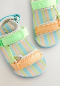 Laste sandaalid pastelse oranži ja rohelise reguleeritava rihmaga ning triibulise sinise, rohelise ja oranži taldaga valgel taustal.